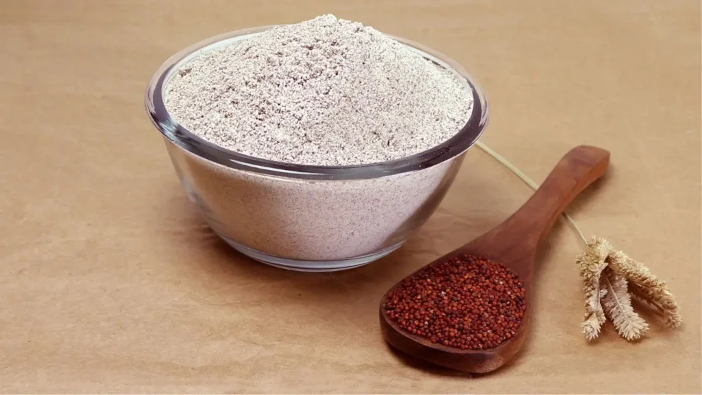 एक कांच के बाउल में रागी का आटा और लकड़ी के चम्मच में साबुत रागी के दाने रखे हुए हैं।