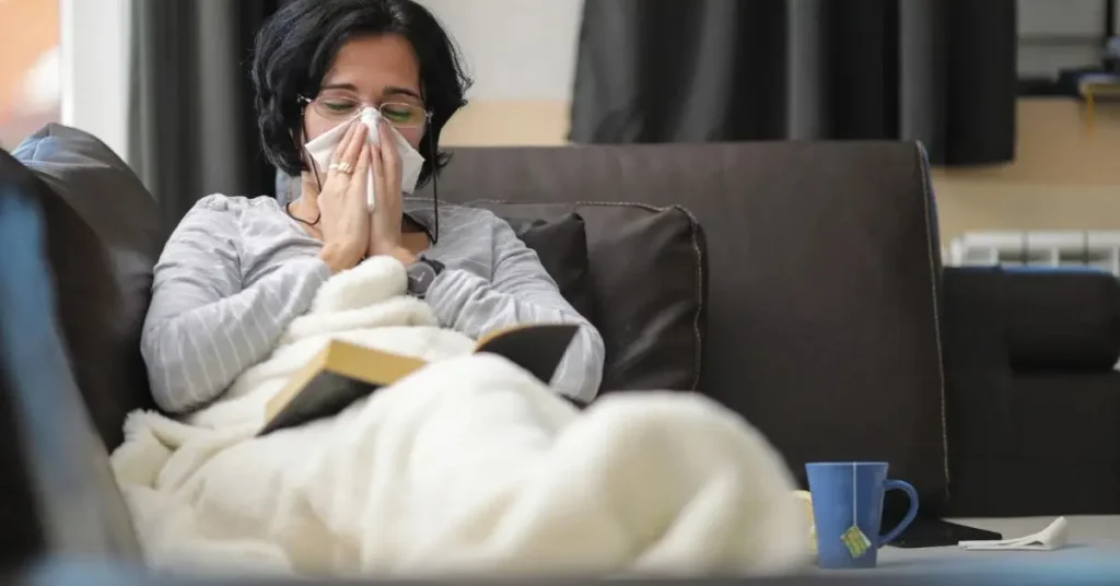 सोफ़े पर बैठी महिला H3N2 फ्लू से पीड़ित, मास्क पहनकर और टिश्यू से नाक पोंछते हुए