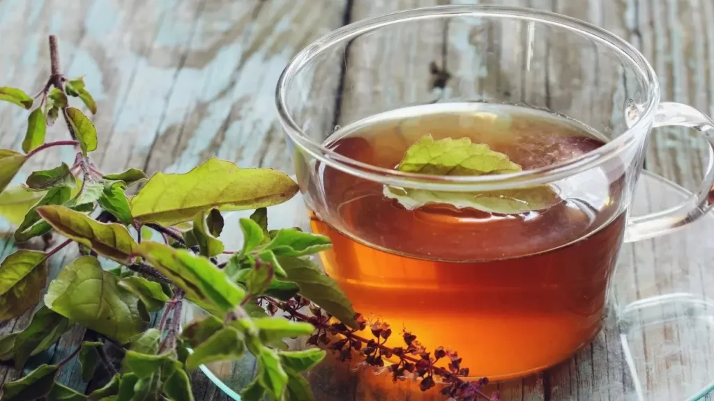 गर्मागर्म तुलसी (tulsi) चाय/उबला तुलसी पानी का कप, ऊपर तुलसी की पत्ती और पास में ताज़ी पत्तियाँ लकड़ी की सतह पर