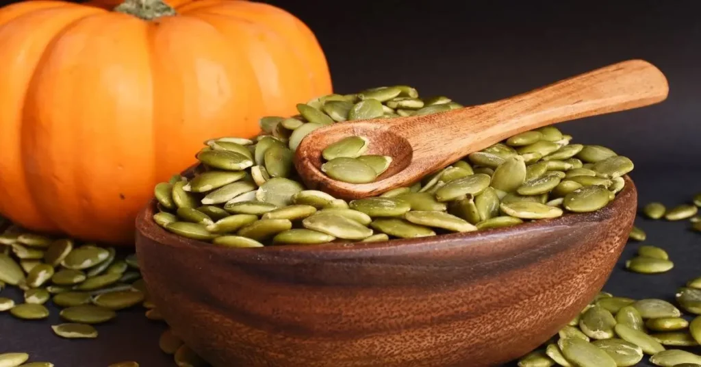 pumpkin seeds से भरा लकड़ी का कटोरा, ऊपर लकड़ी का चम्मच, पीछे पूरा कद्दू, डार्क बैकग्राउंड पर क्लोज़-अप।