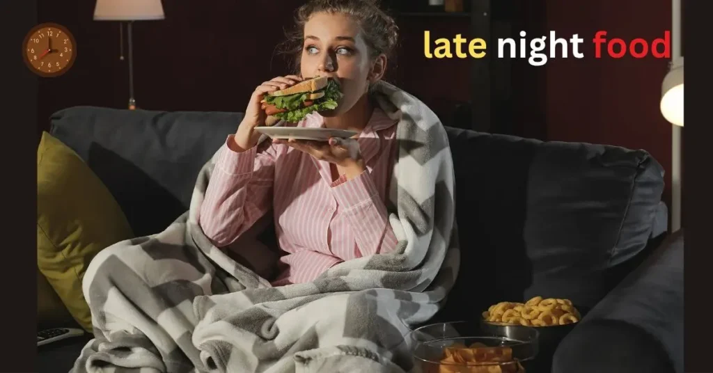 कंबल ओढ़कर सोफ़े पर बैठी युवती देर रात खाना खा रही है—सैंडविच और स्नैक्स के बाउल सामने रखे हैं, कमरे में हल्की रोशनी और दीवार पर घड़ी; लेट-नाइट खाना आदत को दिखाता दृश्य।