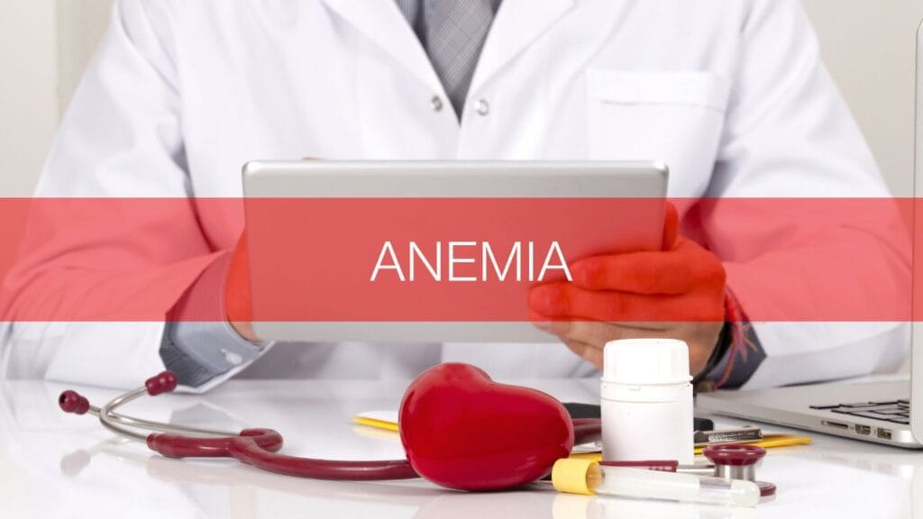डॉक्टर सफेद कोट में टैबलेट पकड़े हुए हैं, स्क्रीन पर “ANEMIA” लिखा है और सामने स्टेथोस्कोप, दवा की बोतल तथा हार्ट मॉडल रखा हुआ है।
