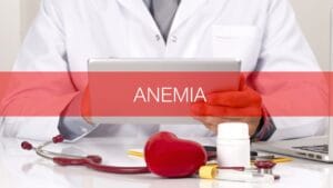 डॉक्टर सफेद कोट में टैबलेट पकड़े हुए हैं, स्क्रीन पर “ANEMIA” लिखा है और सामने स्टेथोस्कोप, दवा की बोतल तथा हार्ट मॉडल रखा हुआ है।